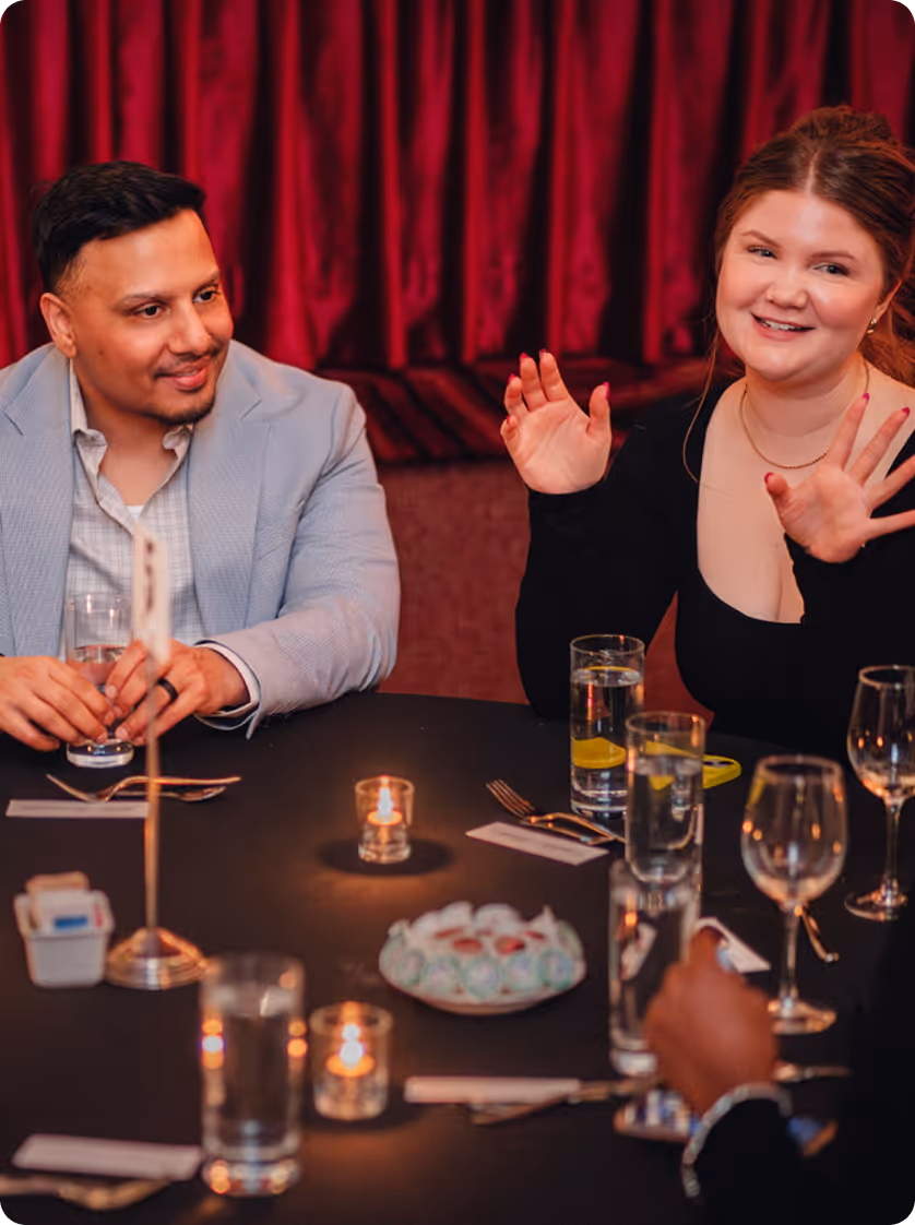 Deux personnes assises à une table dans un environnement faiblement éclairé, une femme souriante faisant des gestes avec ses mains et un homme en veste légère tenant un verre d'eau.