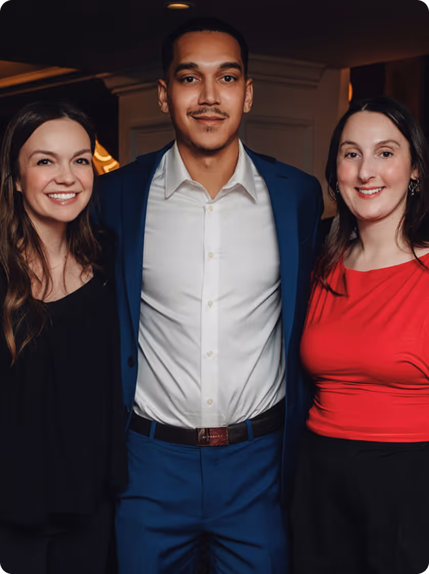 Trois adultes souriants : un homme en costume bleu et chemise blanche se tient entre deux femmes, l'une en haut noir et l'autre en haut rouge.