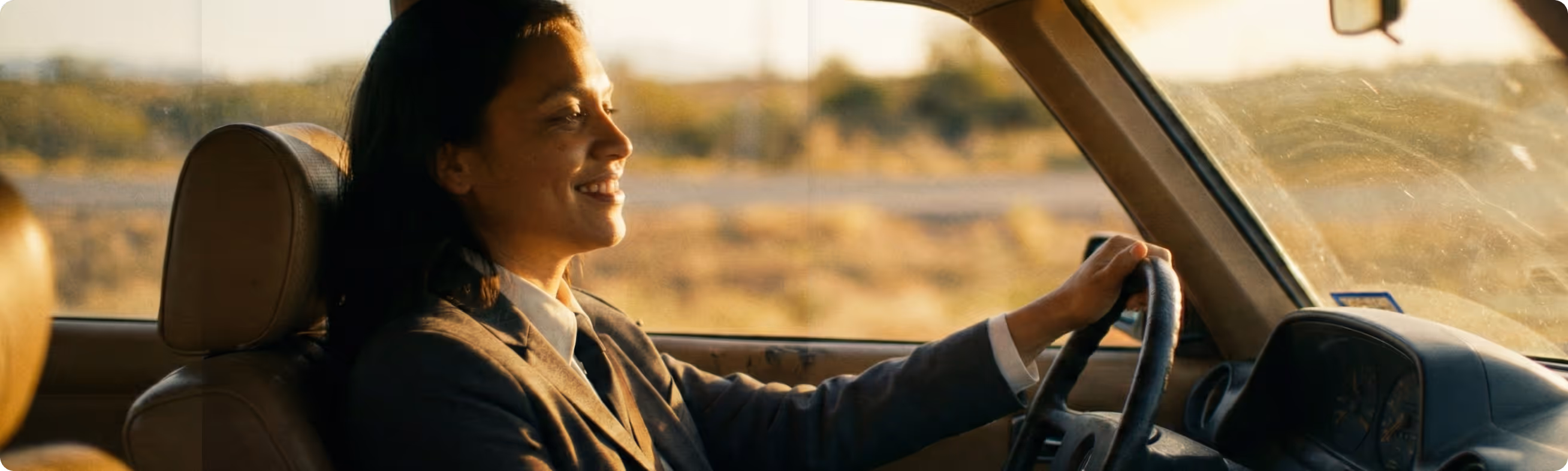 Smiling woman in blazer driving a car during sunset on a country road.