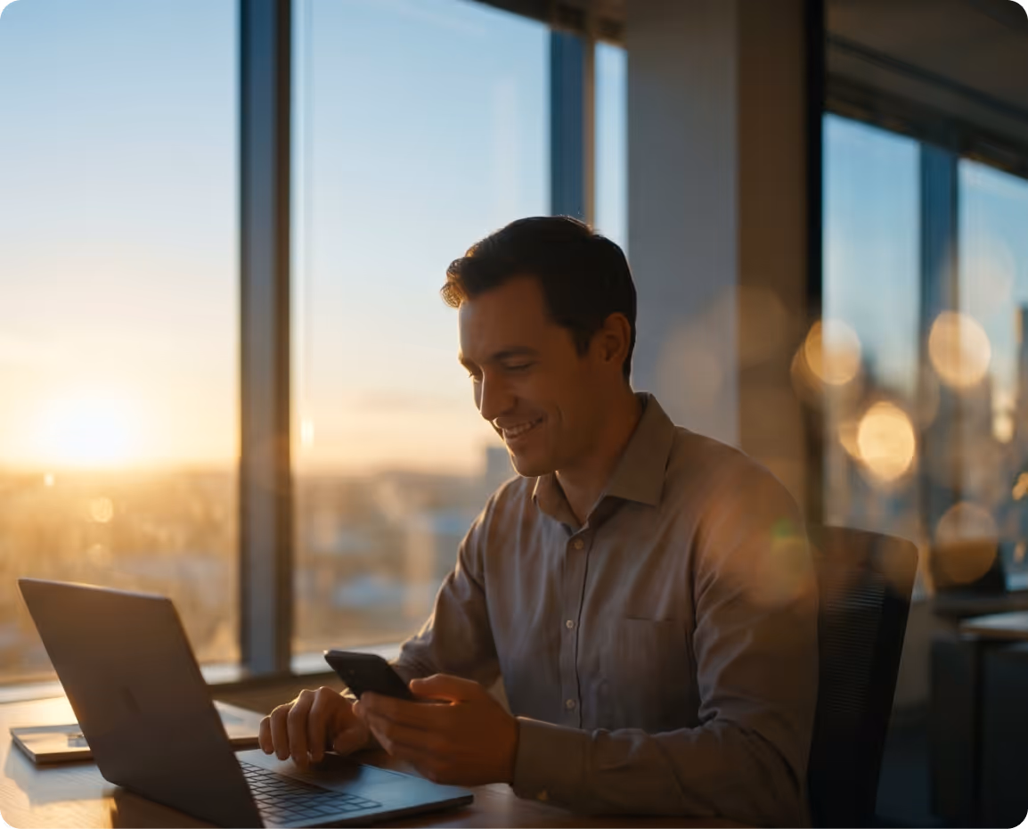 Smiling man sitting at desk using a smartphone with a laptop open during sunset through large office windows.