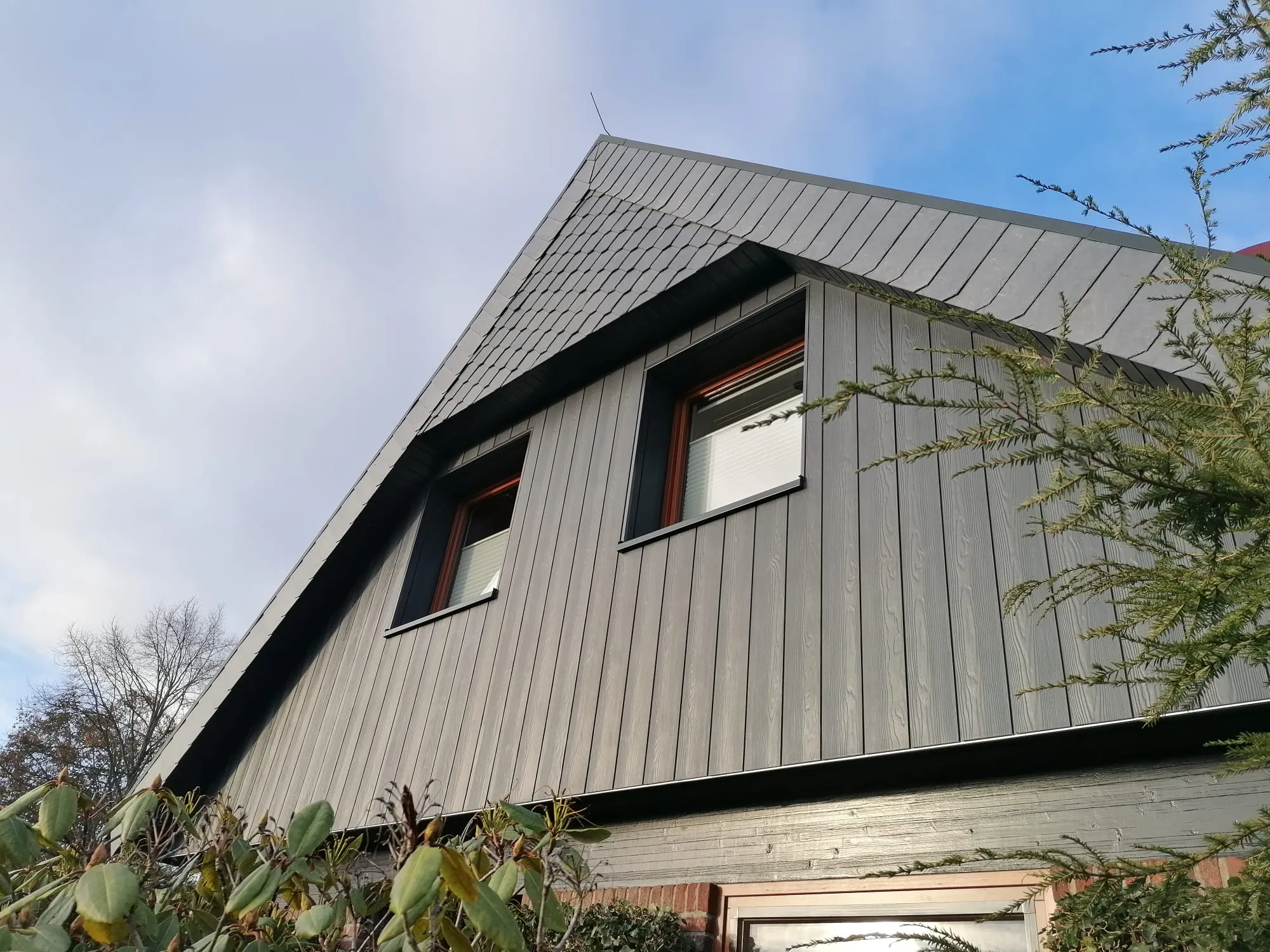 Fassade eines modernen Hauses mit grauer Holzverkleidung und zwei abgedunkelten Fenstern.