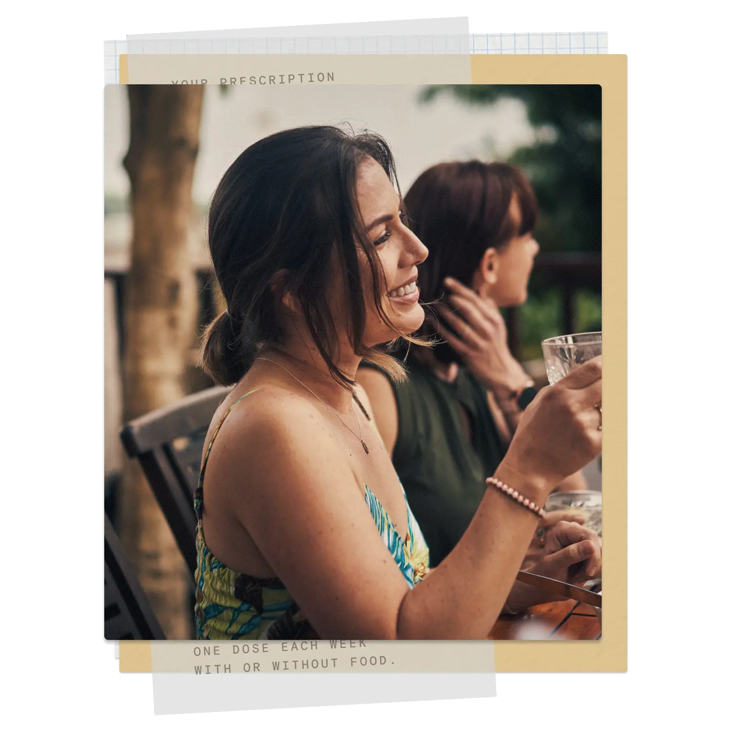 Two women sitting outdoors at a wooden table, one smiling and holding a glass, the other looking away.