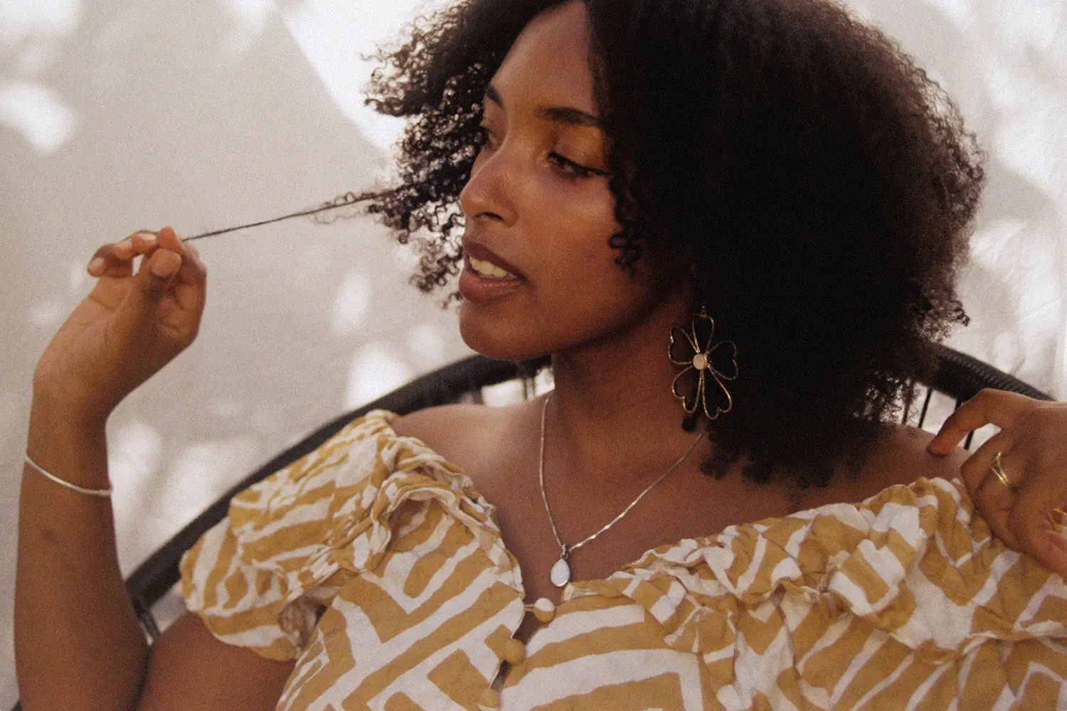 Woman with curly hair wearing a yellow and white patterned off-shoulder top, playing with a strand of her hair.