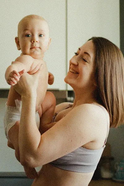 Smiling woman holding a baby wearing a diaper in a bright room.
