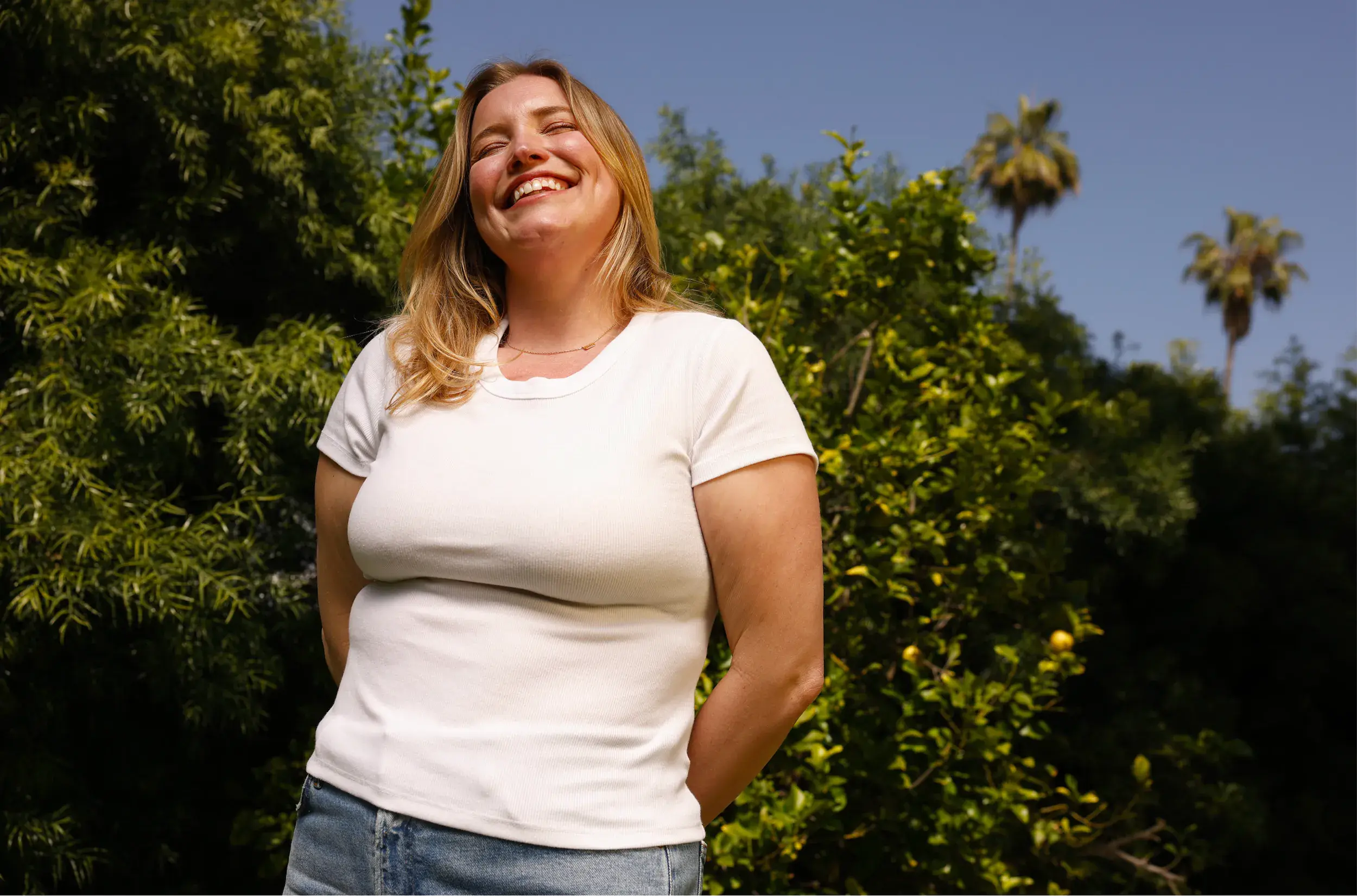 Smiling woman with blonde hair in a white t-shirt and jeans standing outdoors in front of green leafy trees and palm trees under a clear blue sky.