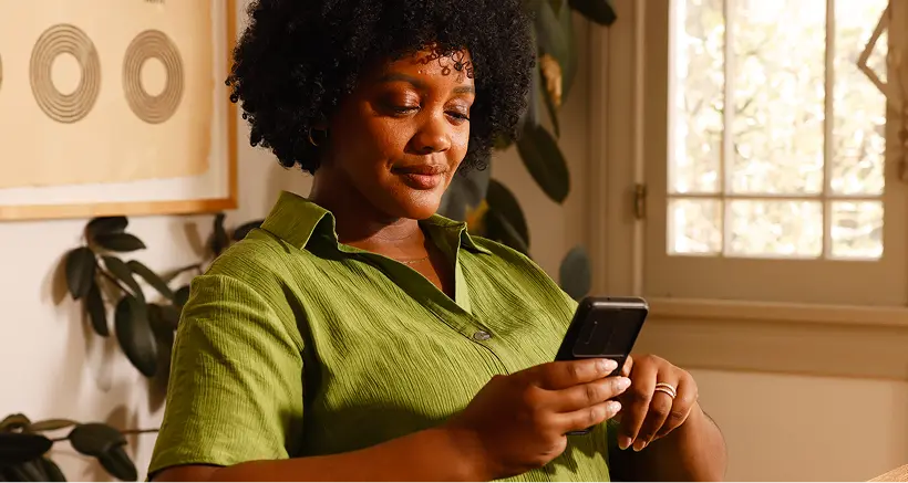 A woman with curly hair wearing a green shirt is sitting indoors, looking at and using her smartphone with a calm expression, near a window and houseplants.