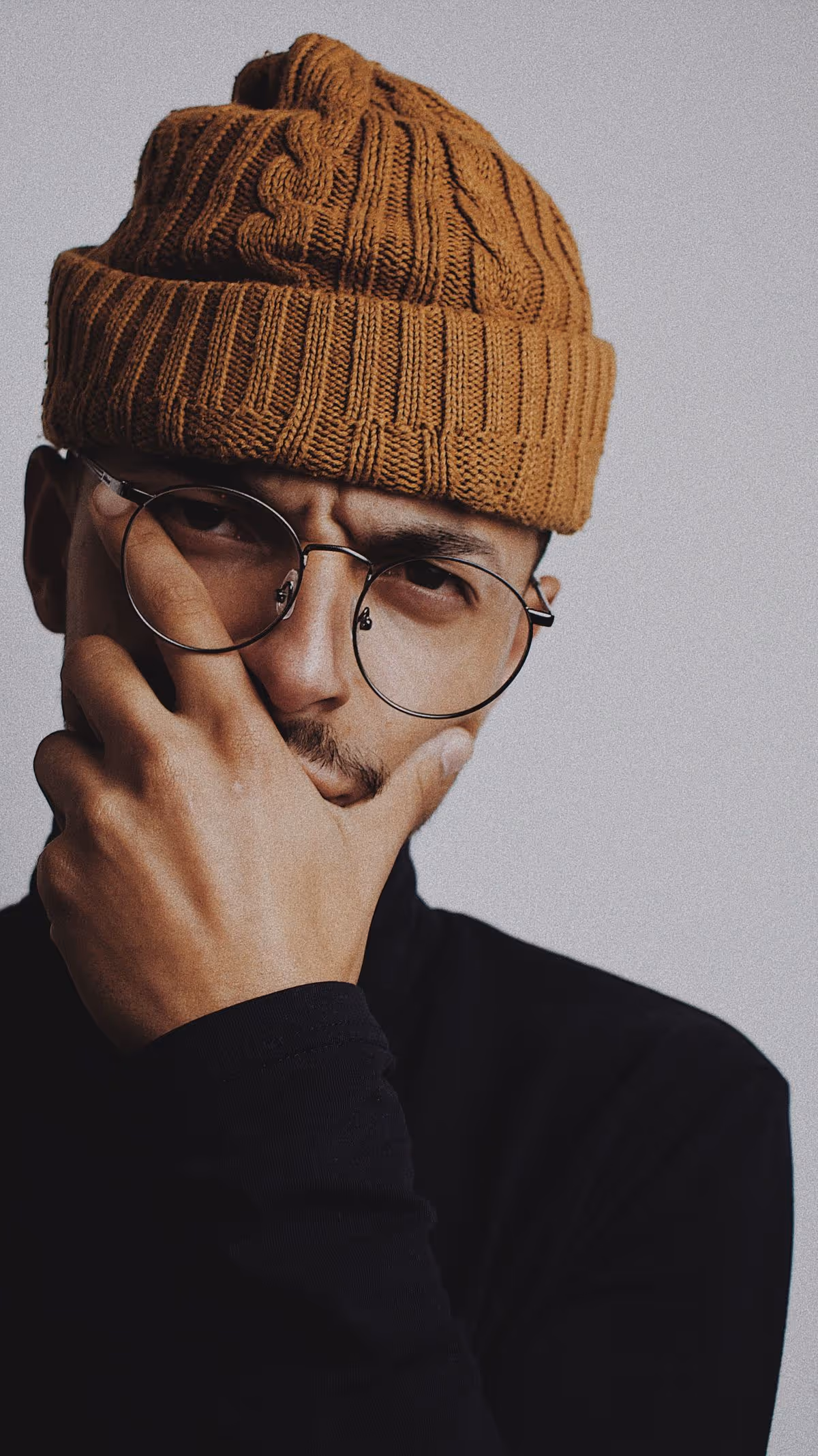 Man wearing round glasses and a brown knit beanie, with his hand partially covering his mouth.