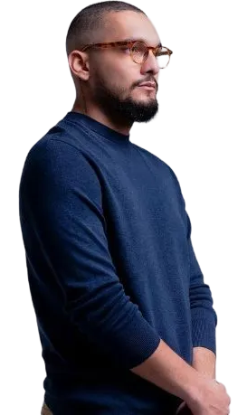 Man with glasses and beard wearing a navy blue sweater, standing with hands clasped, looking to the right.
