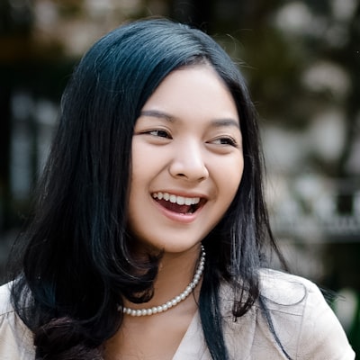 Smiling woman with long black hair wearing a pearl necklace and light-colored top outdoors.