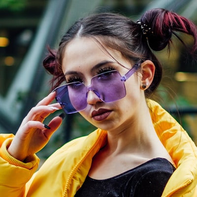 Young woman wearing purple-tinted sunglasses and a yellow jacket with hair styled in two buns.