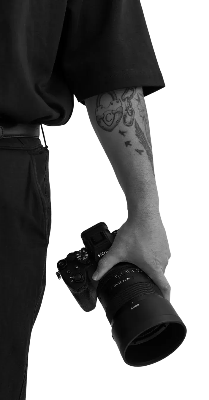 Person wearing black clothing holding a Sony camera with a large lens and showing tattoos on their forearm.