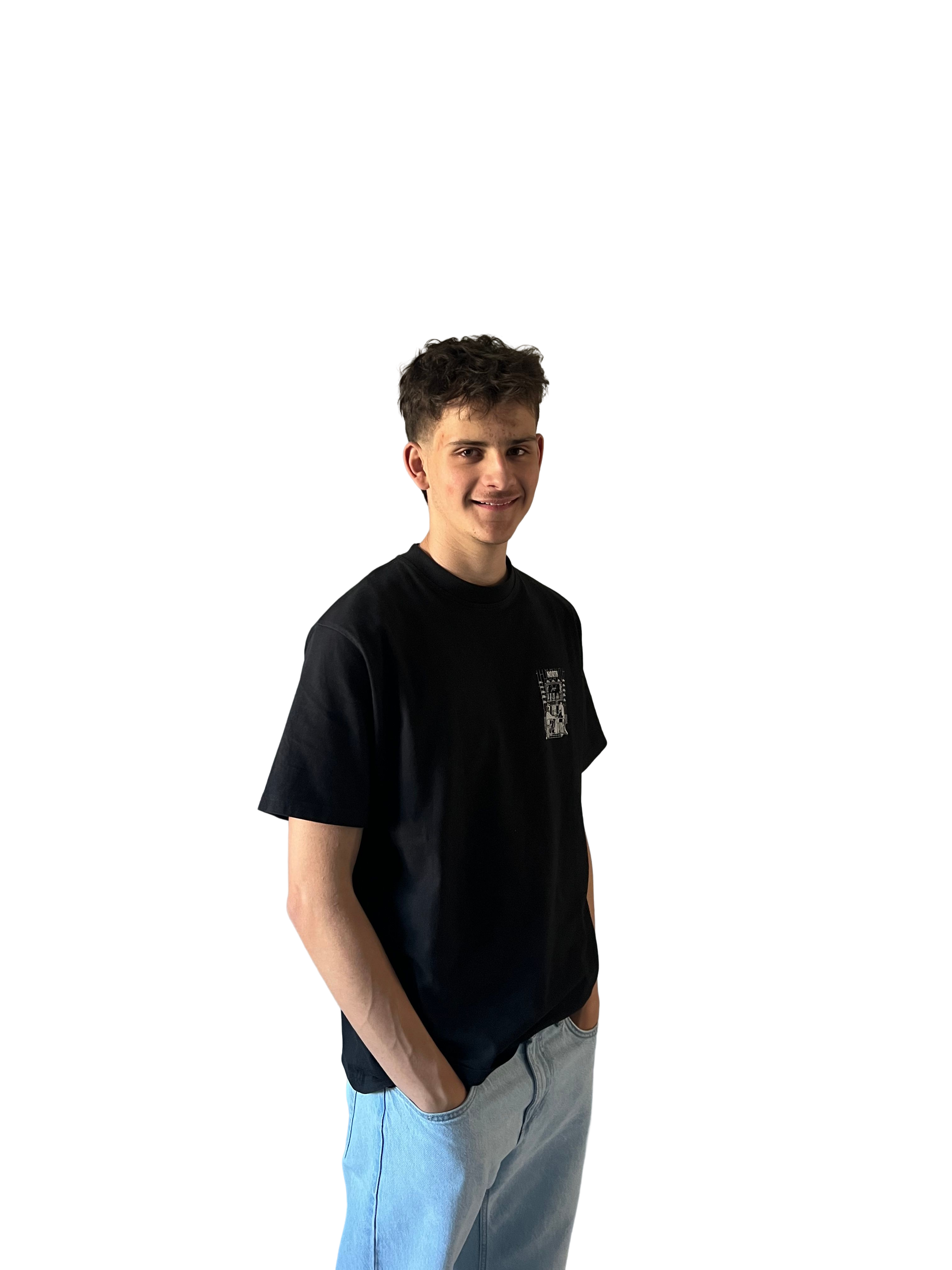 Young man with curly dark hair wearing a black t-shirt and light blue jeans, standing with hands in pockets and smiling.
