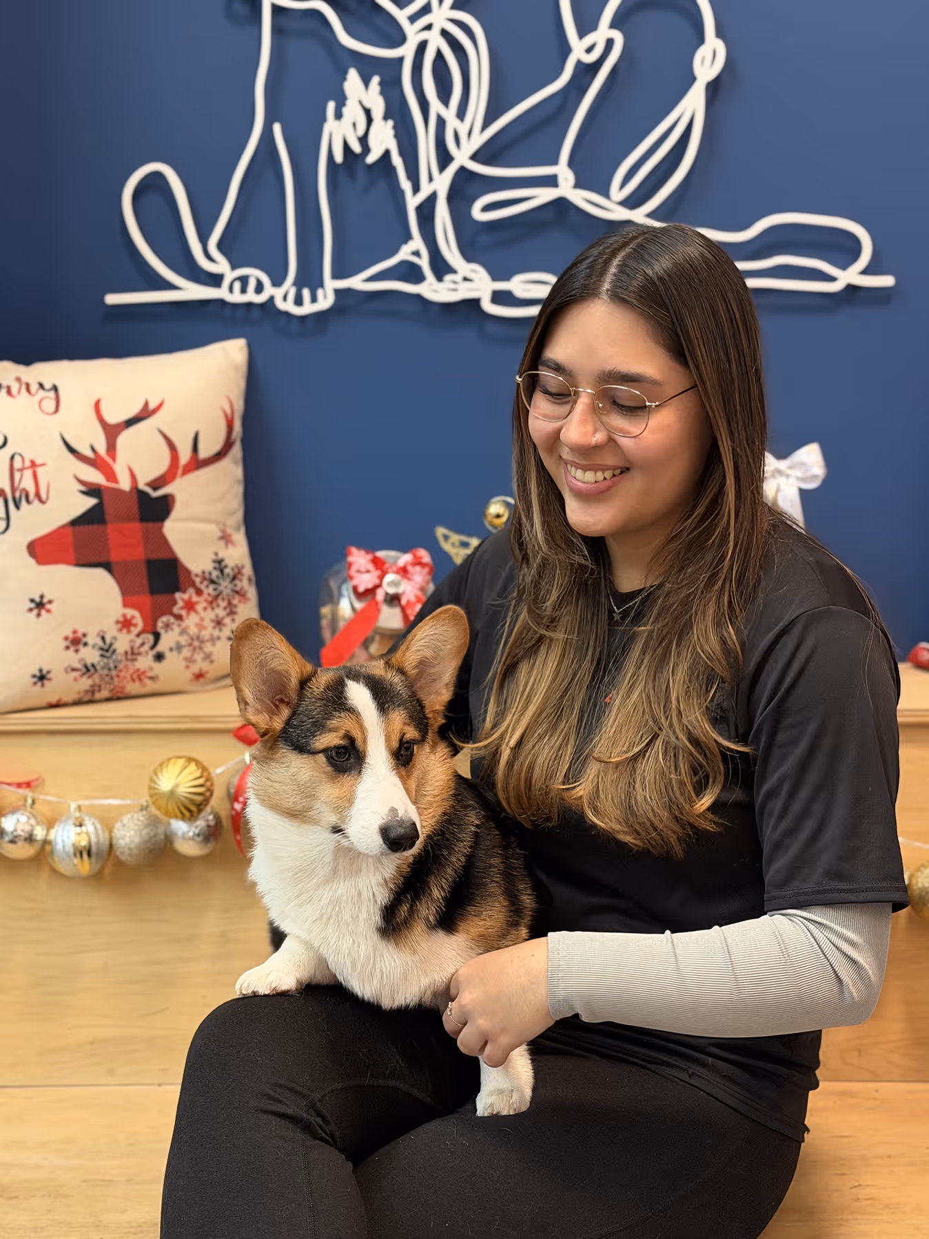 Smiling woman with glasses holding a tricolor corgi dog, festive holiday decorations and a reindeer pillow in background.