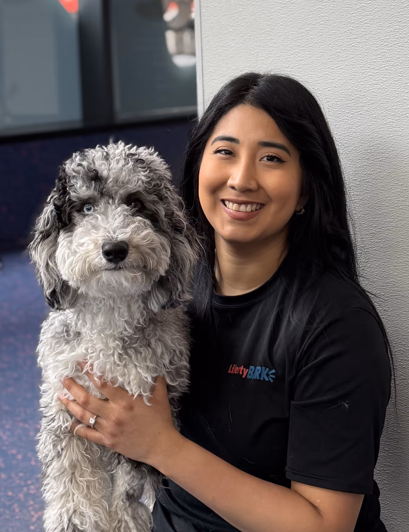 Smiling woman with long black hair holding a curly gray and black dog with one blue eye indoors.