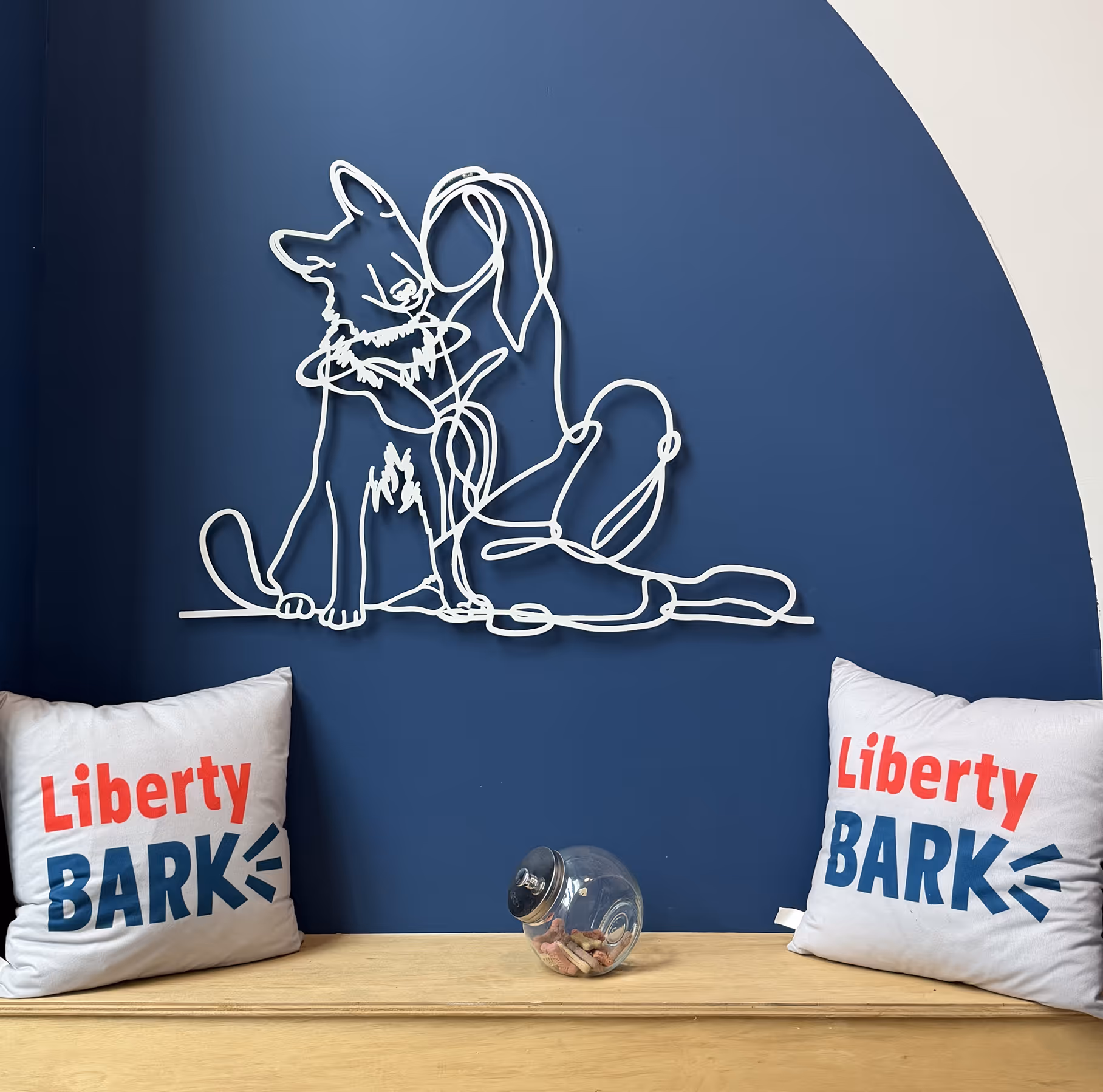 White wire art of a woman hugging a dog on a blue wall with two pillows labeled 'Liberty BARK' and a wooden surface with a tilted glass jar of dog treats.