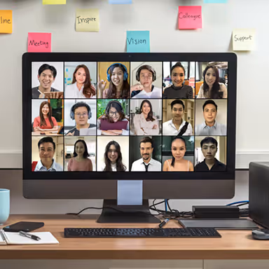Desktop computer on a desk showing a video conference with 20 diverse participants arranged in a grid, surrounded by sticky notes with motivational words.