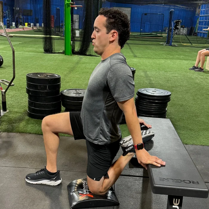 Man performing a kneeling hip flexor stretch on a padded mat while supporting himself with hands on a bench in a gym.