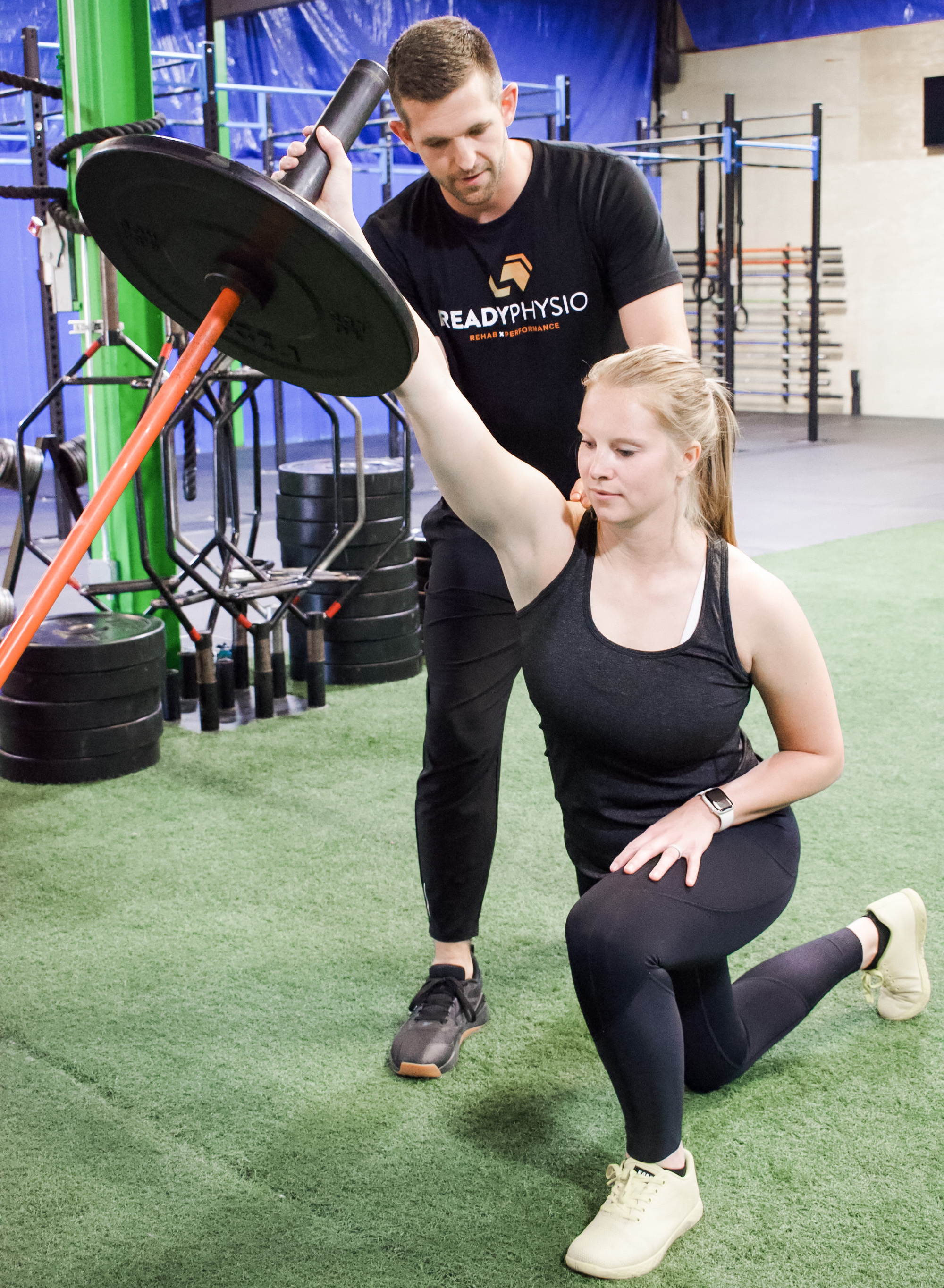 A woman in black workout clothes performs a kneeling landmine press with a weighted bar, assisted by a male trainer in a Ready Physio shirt inside a gym.