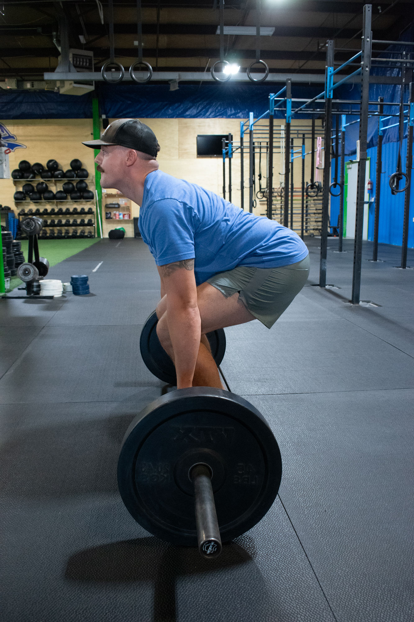 Man in a blue shirt and shorts lifting a barbell in a gym with racks and gymnastic rings.