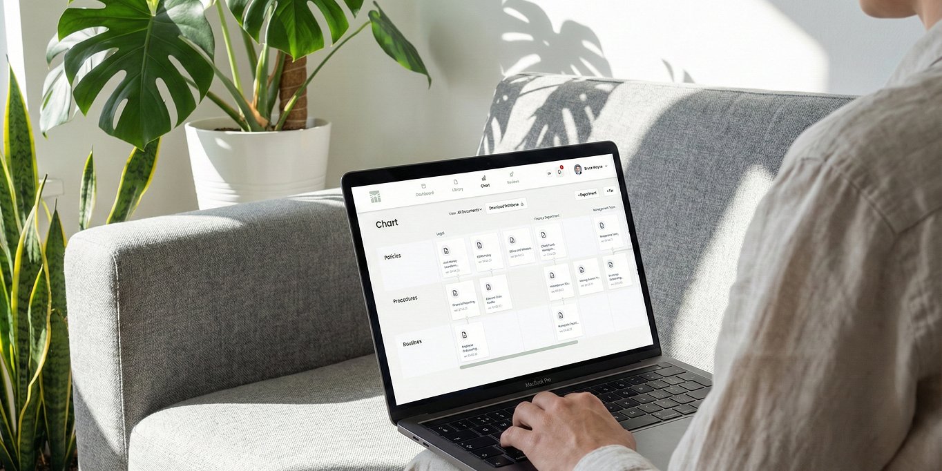 Person using a laptop on a grey sofa with a plant in the background, viewing a document management chart on screen.