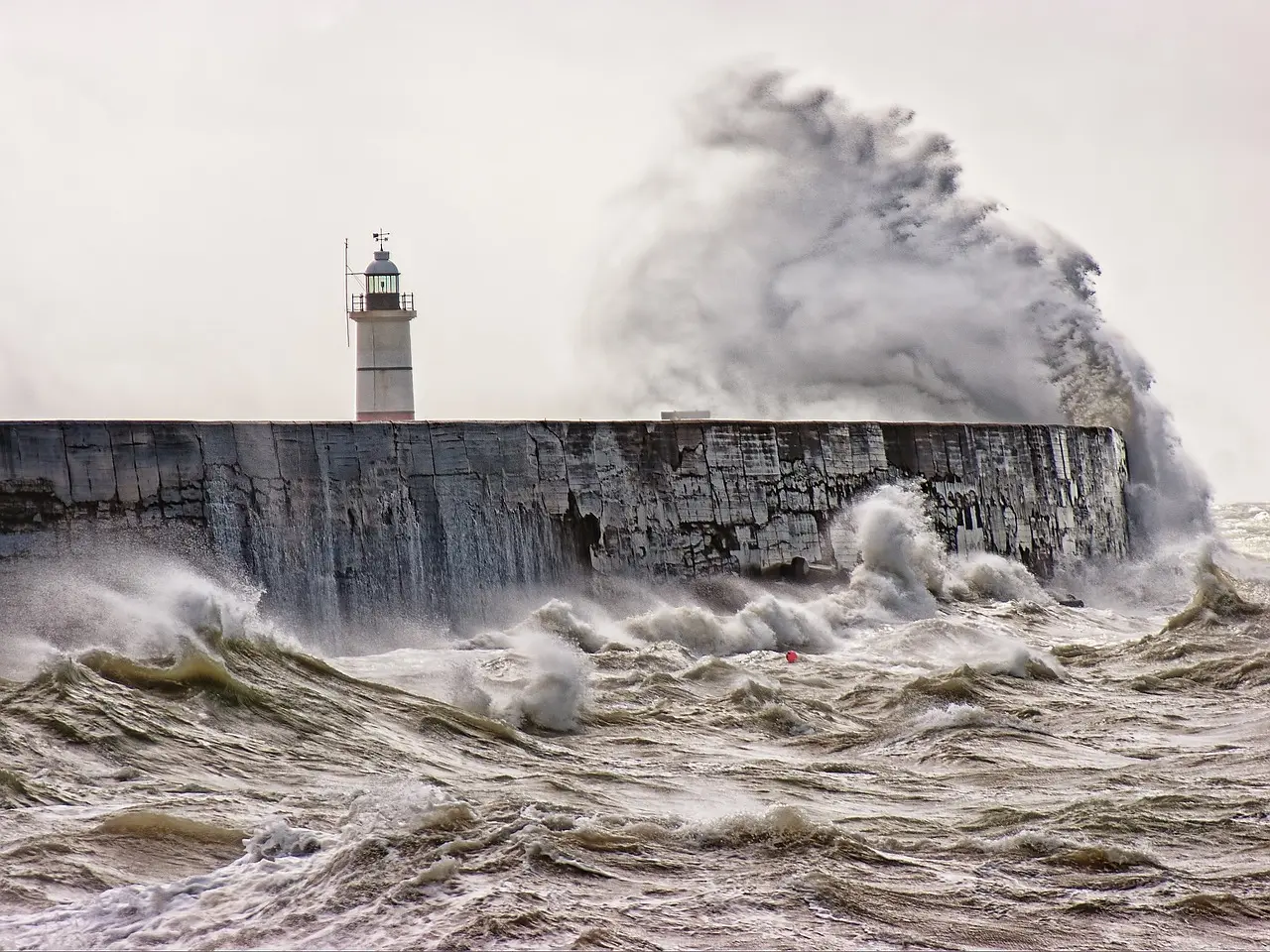 Stormy sea with large waves crashing against a stone breakwater and a white lighthouse.