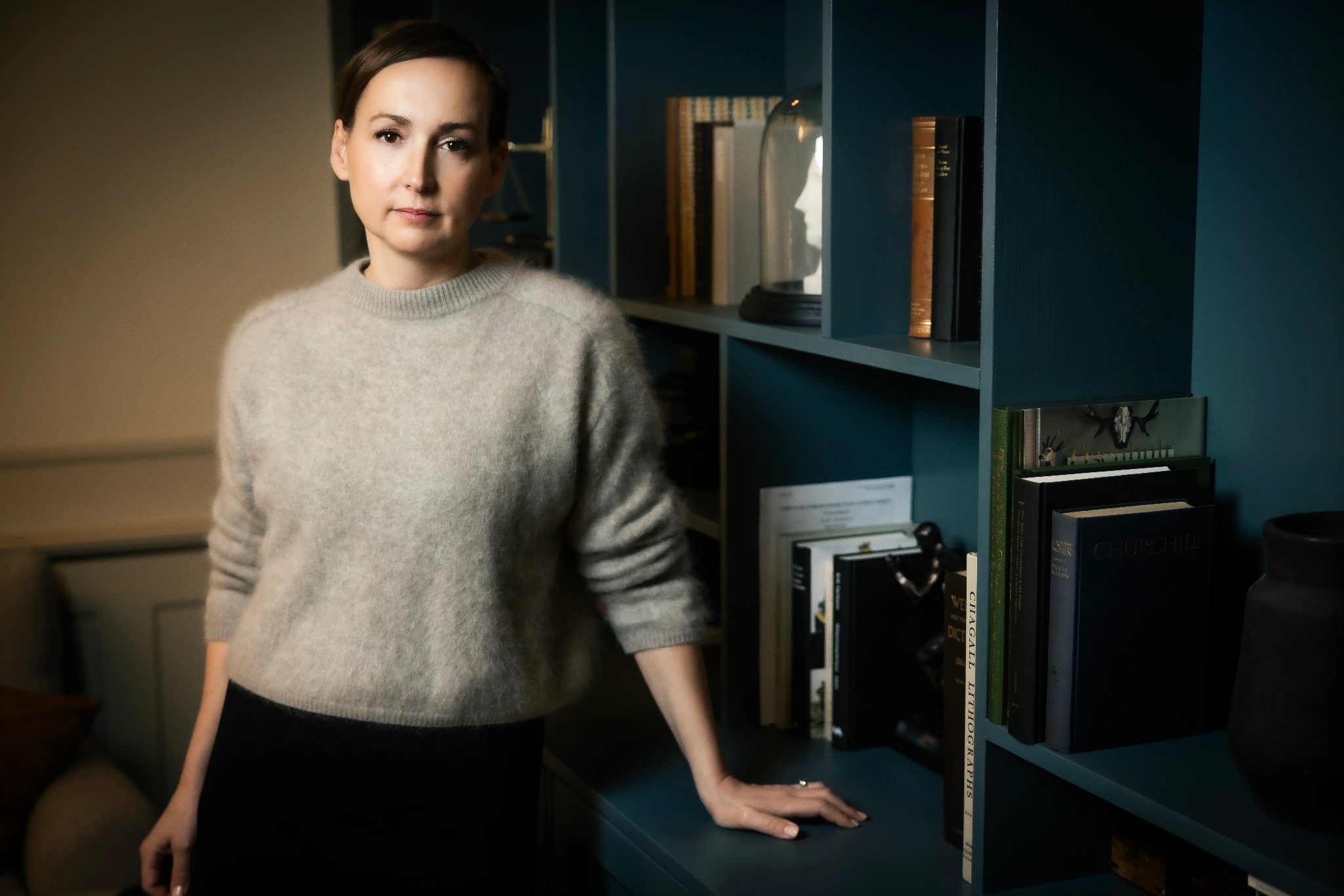 Woman with brown hair wearing a gray sweater leaning on a dark blue bookshelf filled with books and decorative items.