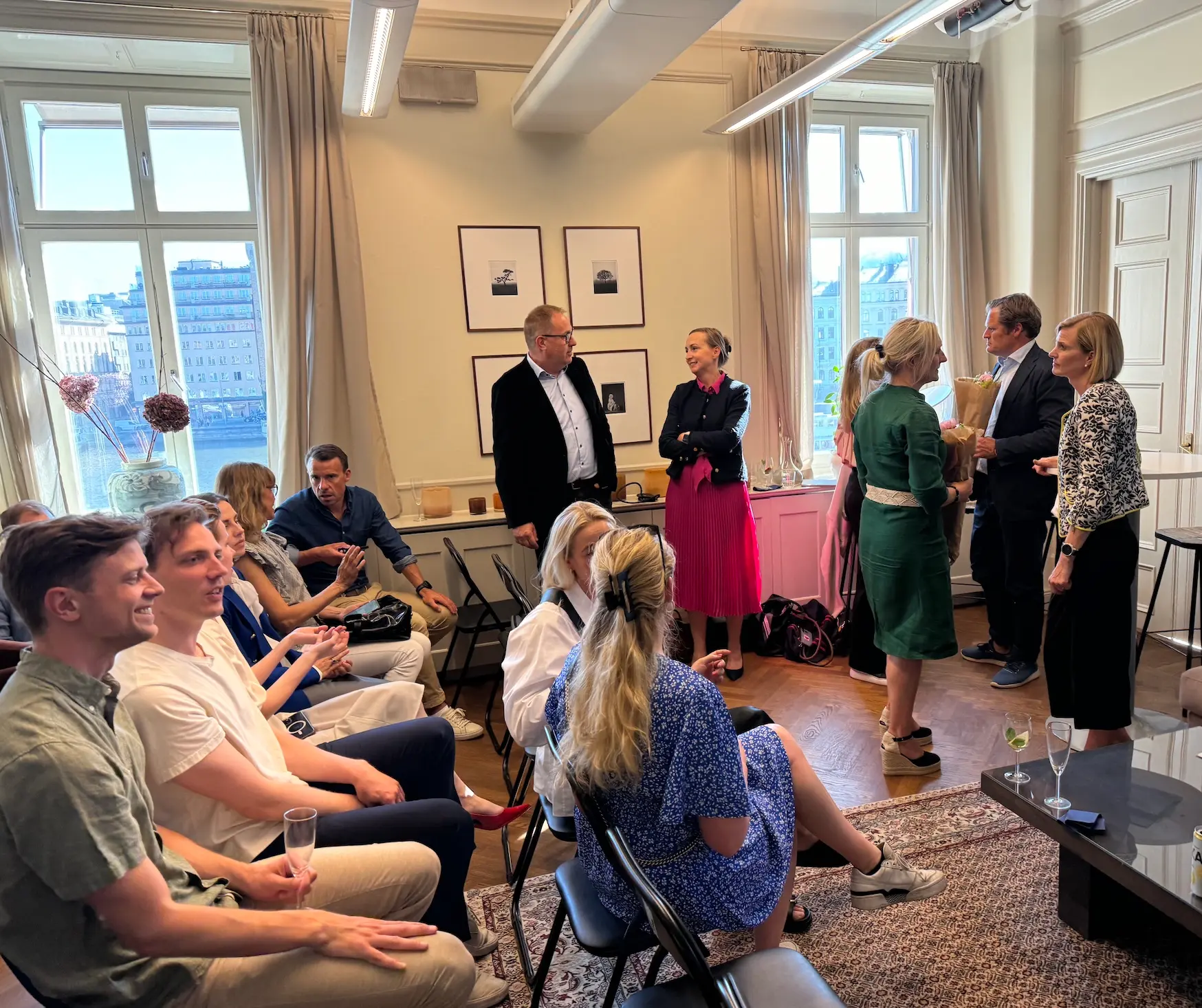 Group of people seated and standing in a bright room with large windows, engaged in conversation during a social gathering.