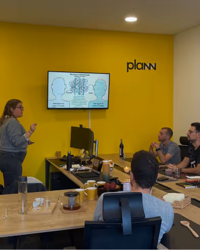 PLANN team member presenting to three colleagues in a modern office with a yellow wall and Plann logo.