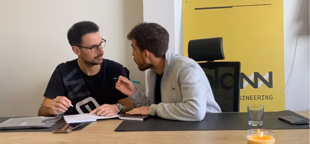 Two PLANN team members having a discussion over documents at a desk with a laptop, glass, and phone nearby.