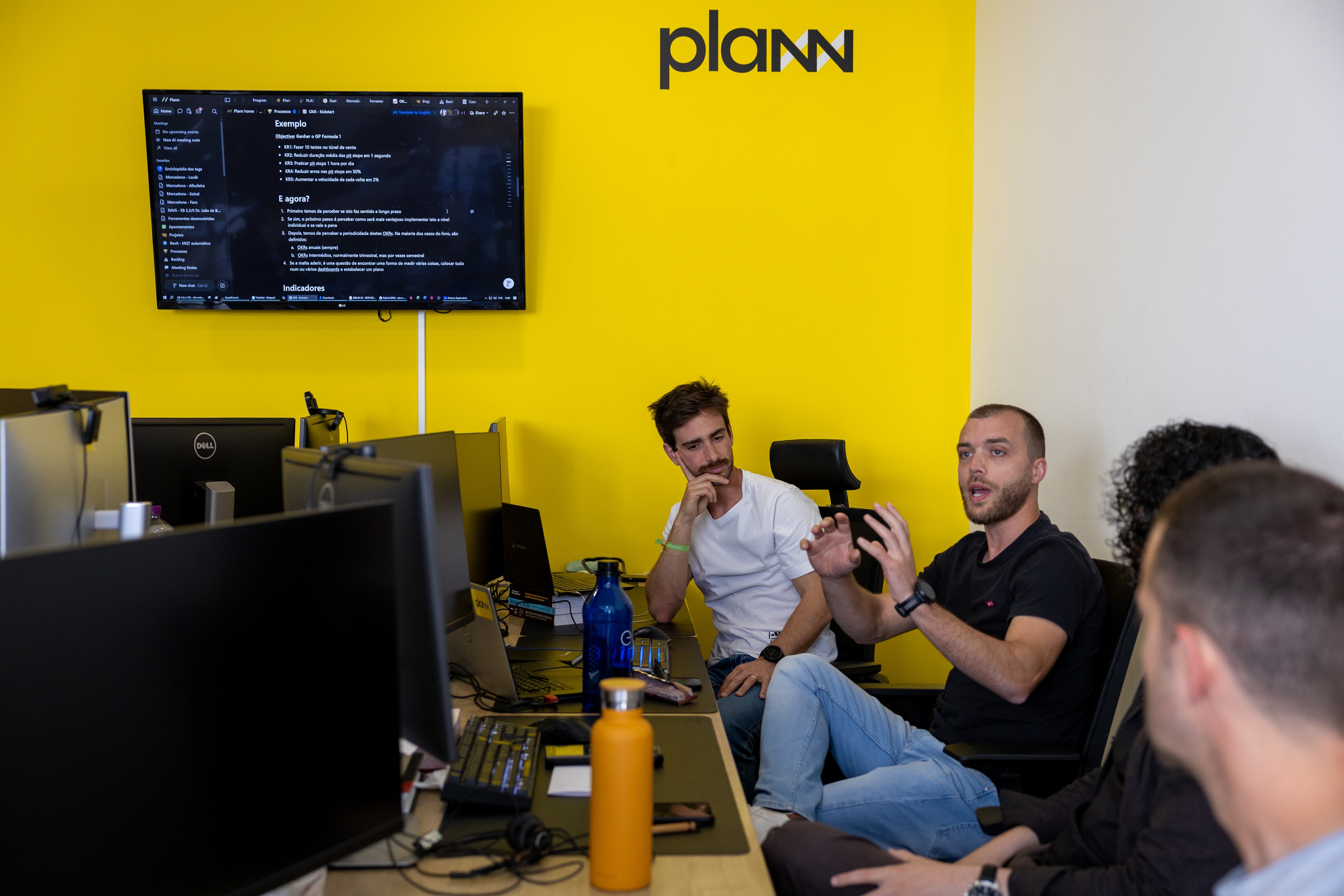 Four people in an office having a discussion; one man in a black t-shirt is speaking with hand gestures, while others listen attentively; a yellow wall with the logo 'planW' and a mounted screen displaying text are in the background; desks with multiple computer monitors and water bottles are visible in the foreground.