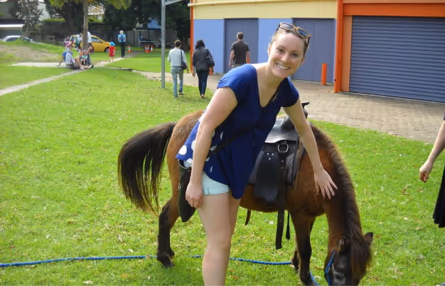 Smiling woman in blue shirt and white shorts petting a brown pony with a saddle on a grassy area with people walking in the background.