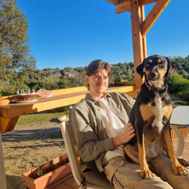 A young man sitting outdoors on a chair with a black and tan dog on his lap, under a wooden pergola on a sunny day.