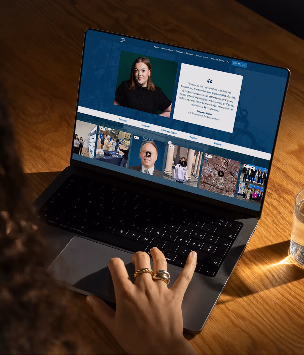 Person browsing the George Washington University website on a laptop, showing a testimonial with a woman's portrait and various social media posts.