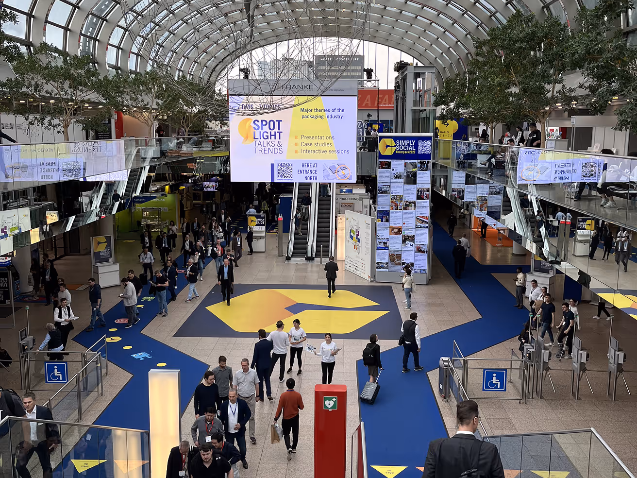 Busy modern exhibition hall with a curved glass ceiling, blue carpet pathways, and a large signboard displaying event information about packaging industry talks.