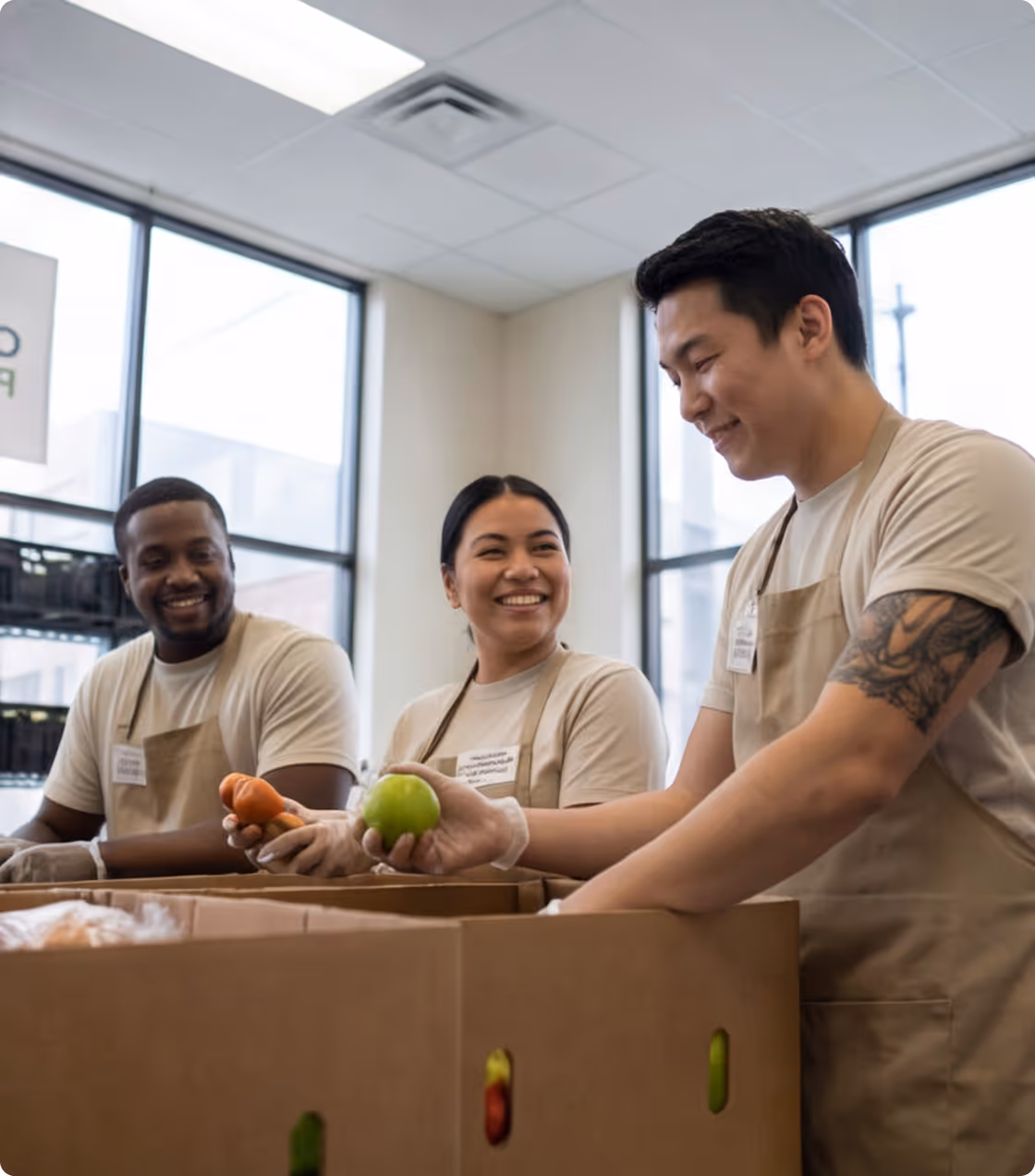 Three diverse volunteers smiling and packing fresh produce into large cardboard boxes indoors.