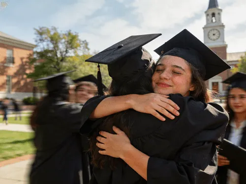 Two graduates in caps and gowns hugging each other with a blurred group of graduates and a clock tower in the background.