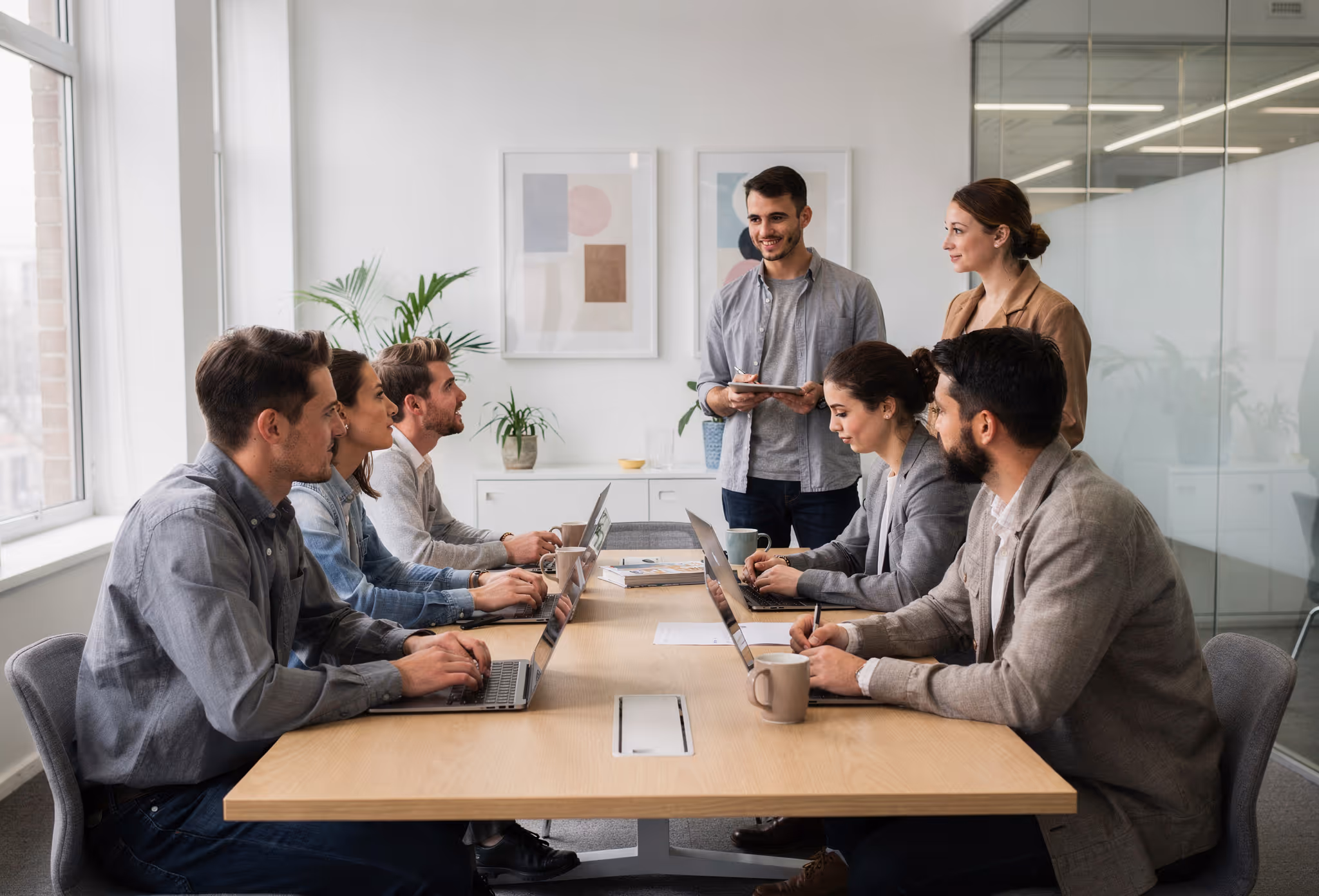 Six colleagues engaged in a meeting around a table with laptops, with two standing presenting in a modern office.