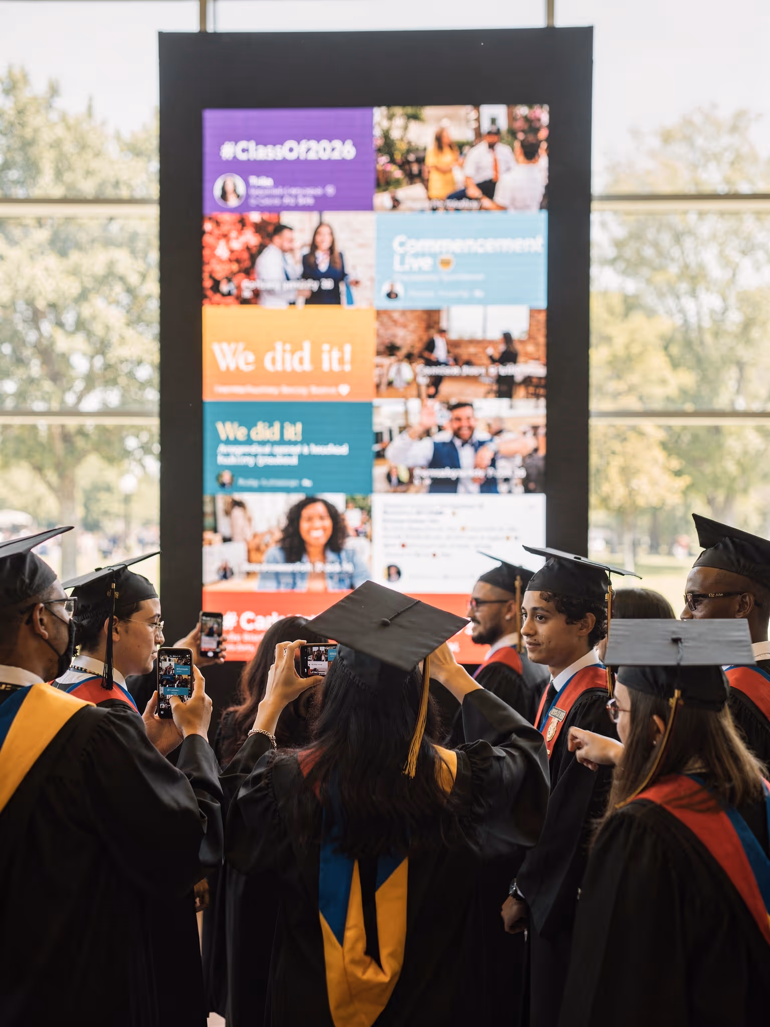 Group of graduates in caps and gowns taking photos with their phones in front of a large screen displaying congratulatory social media posts.