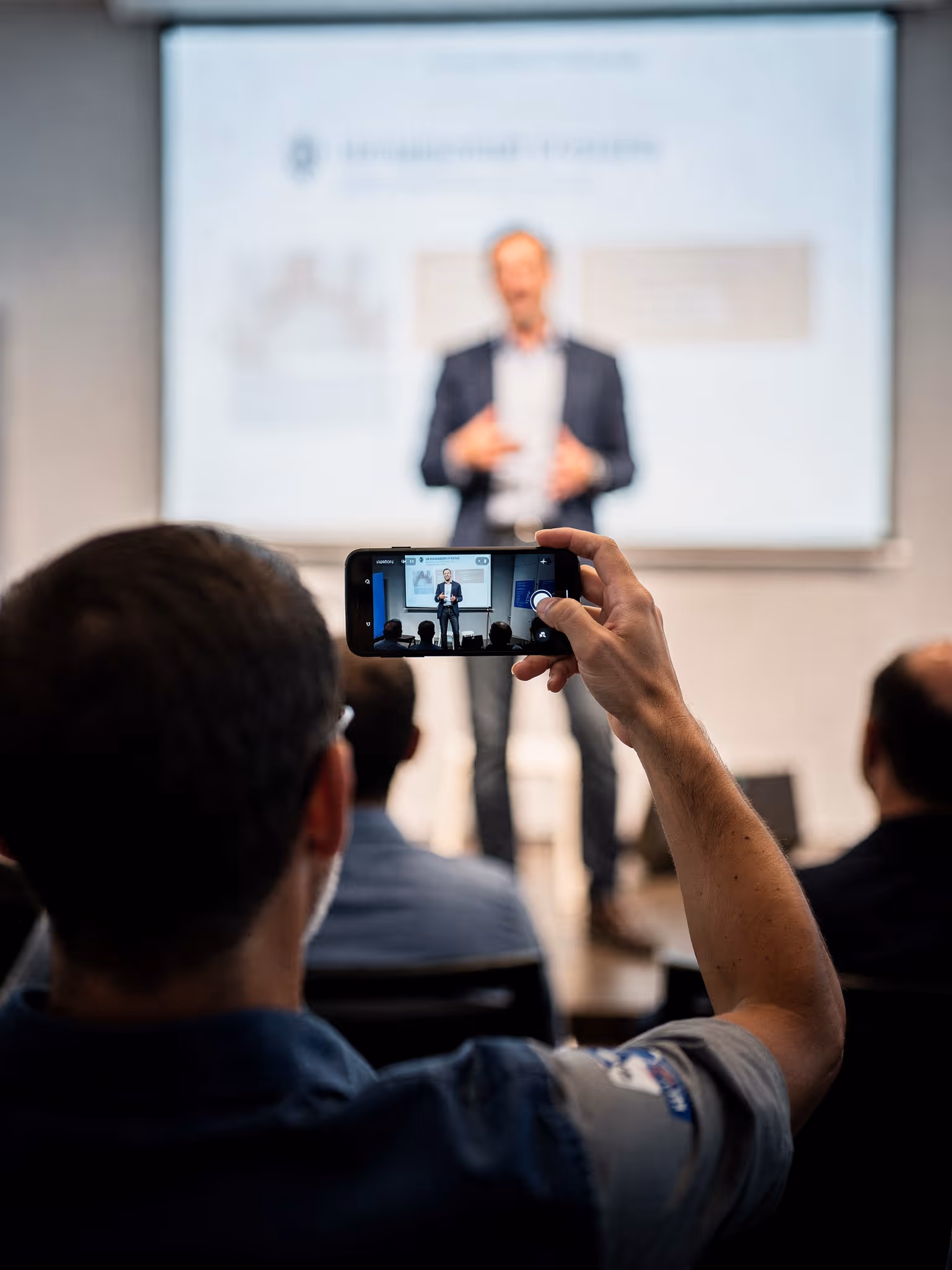 Person recording a speaker giving a presentation on a projector screen with a smartphone during a seminar.