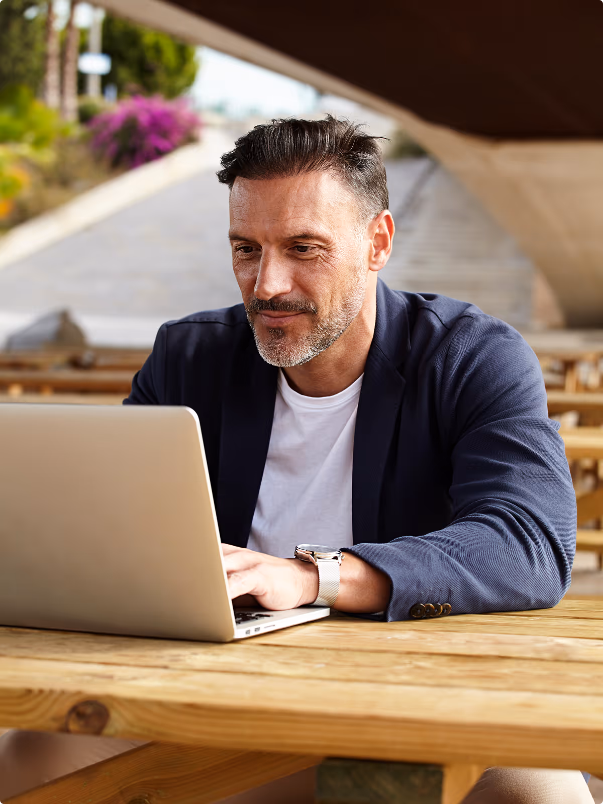 Man in a navy blazer and white shirt working on a laptop at a wooden picnic table outdoors.
