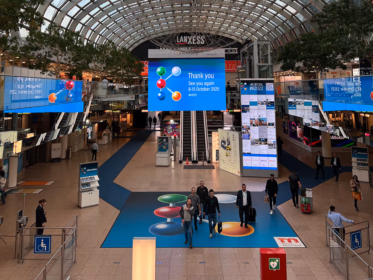 Interior of a large convention center with exhibition booths, escalators, and a digital screen displaying event messaging as attendees walk through the hall.