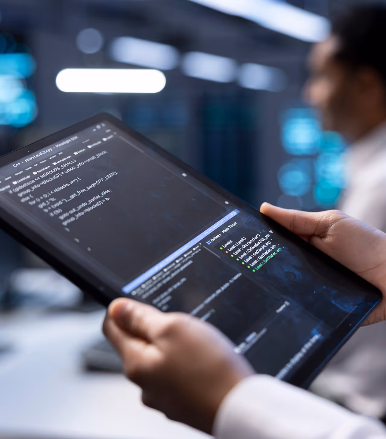 Close-up of an engineer holding a digital tablet displaying lines of C++ code and software development tools in a blurred high-tech laboratory background