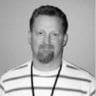 Man with short hair and beard wearing striped shirt and black lanyard, smiling slightly in grayscale photo.