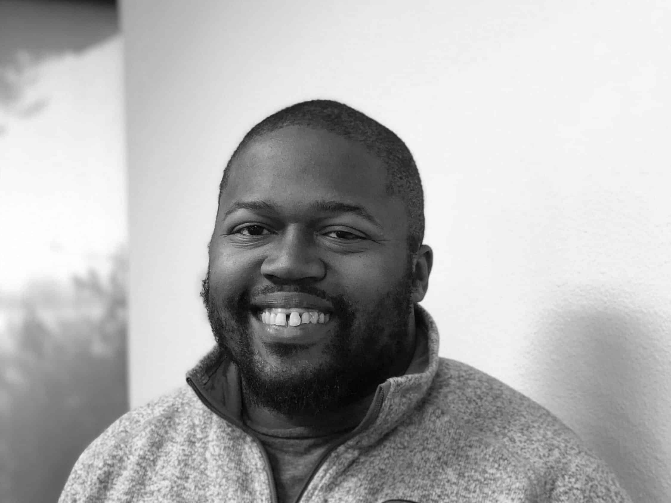Smiling man with short hair and beard wearing a light zip-up sweater against a plain wall.