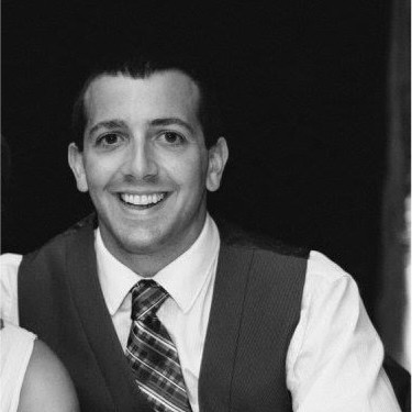 Smiling man in a white shirt, patterned tie, and sleeveless sweater vest against dark background.