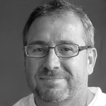 Middle-aged man with glasses, short hair, and beard smiling against gray background.
