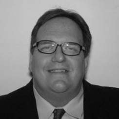 Smiling man wearing glasses, a suit, and a tie against a plain background.