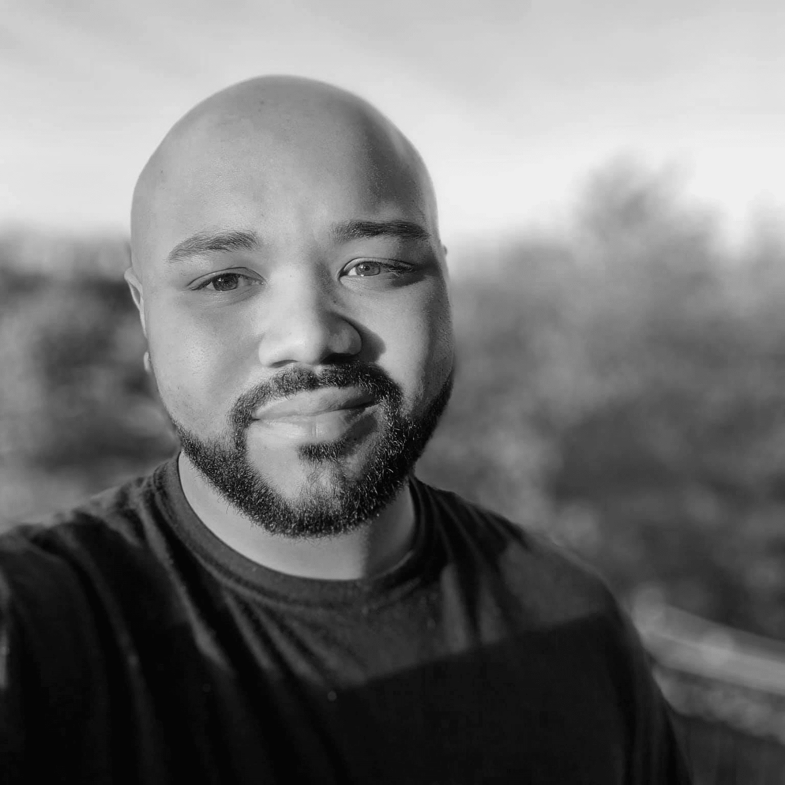 Black and white close-up of a bald man with a beard wearing a dark shirt, smiling slightly outdoors.