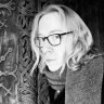 Person with glasses and light hair wearing a dark jacket in front of a textured wall.