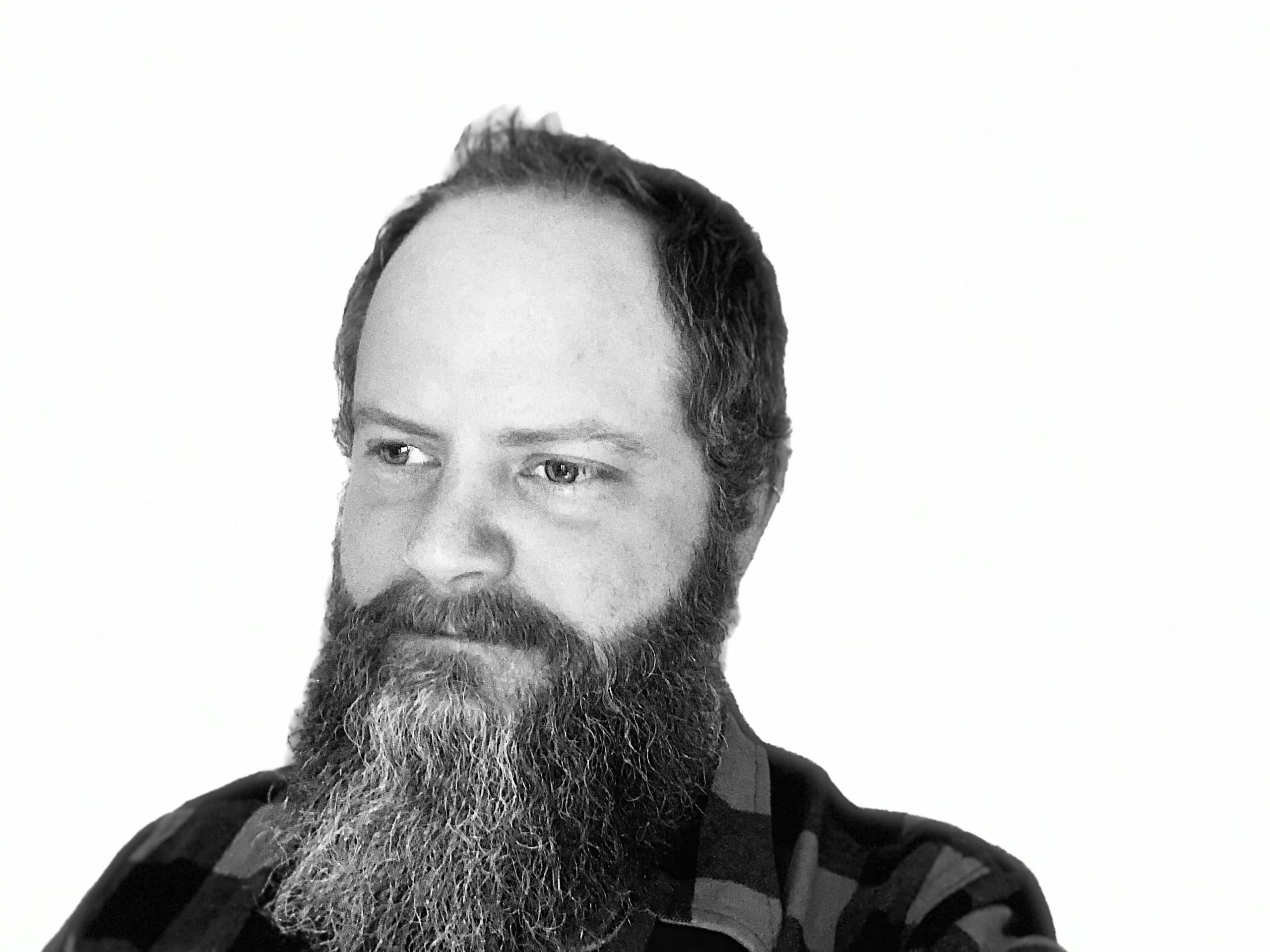 Bearded man with curly hair, wearing a checkered shirt, looking to his right on a white background.